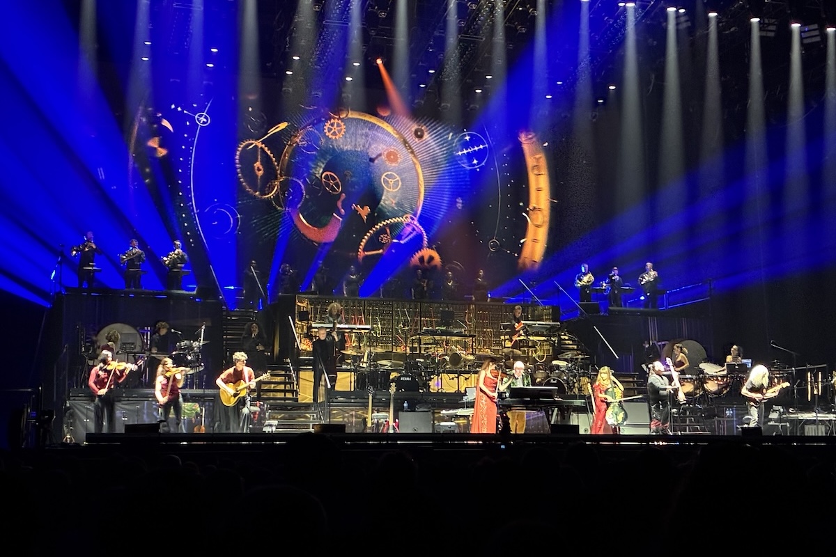 Composer Hans Zimmer performing live on stage with a full orchestra under blue spotlights and cosmic clockwork visuals.